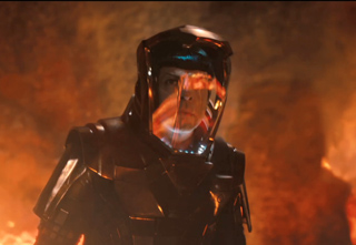 A person wearing dark, heavy protective armor and a large helmet with a clear visor amidst flames.