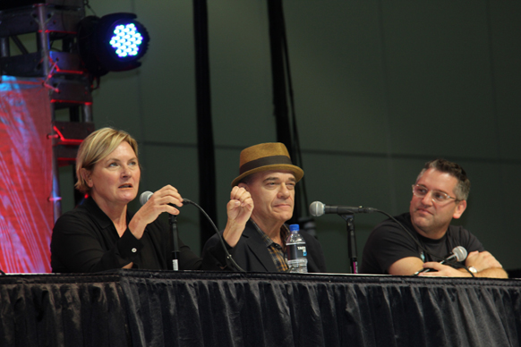Three people sit behind a table with microphones. A woman on the left speaks into a microphone. A man in a hat is in the midd