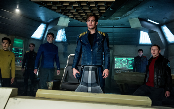 Crew members in blue, yellow, and red uniforms stand on a dimly lit starship bridge.