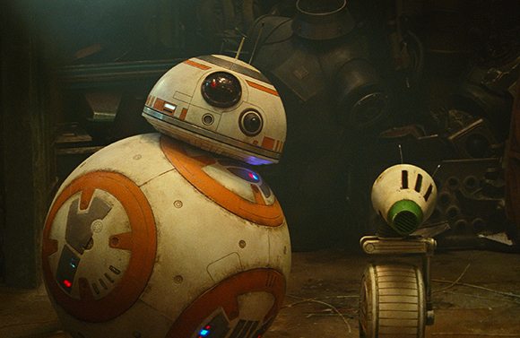 A large white and orange spherical droid stands next to a smaller white droid in a dark, cluttered setting.