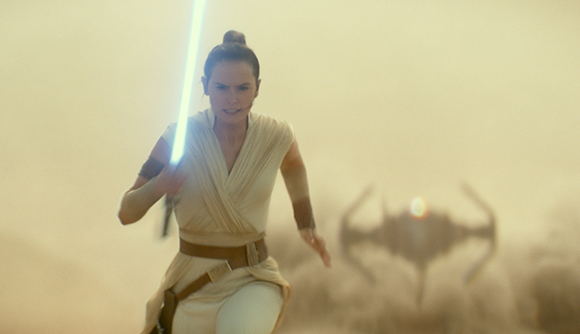 A woman in tan clothing runs through a dusty desert holding a glowing blue lightsaber.