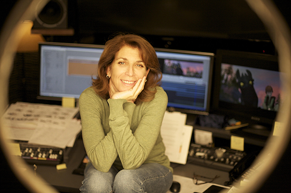 A smiling woman in a green sweater sits in a studio surrounded by computer monitors and editing equipment.