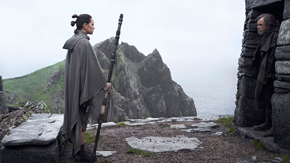 A woman with her hair in buns and holding a staff stands on a rocky cliff, facing a man in dark robes standing in a stone str