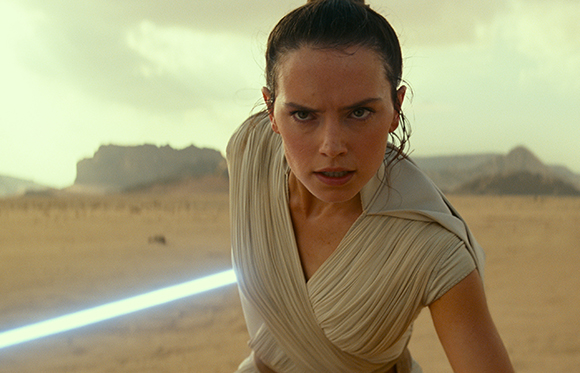 A woman with dark hair pulled back stares intensely while holding a glowing blue lightsaber in a desert.