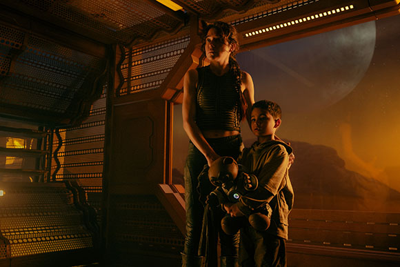 A woman and a young boy stand in a dimly lit spacecraft looking out a window at a large planet.