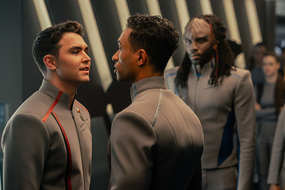 Two men in grey uniforms stand face-to-face in a futuristic hallway while a third man watches from behind.