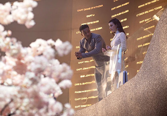 A man and a woman stand before a large wall inscribed with many names.