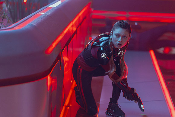 A person in a black and red tactical suit crouches in a futuristic hallway lit by red neon lights.