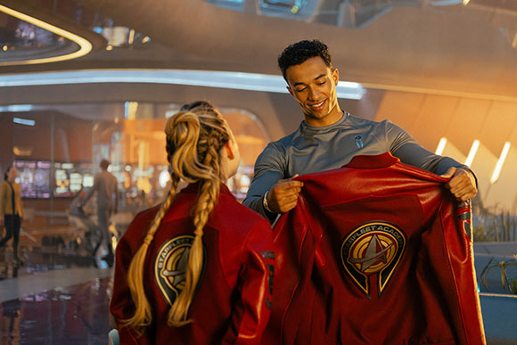 A man smiles while holding up a red Starfleet Academy jacket for a blonde woman.