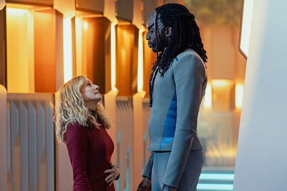 A tall man with long dreadlocks looks down at a blonde woman in a red dress in a futuristic hallway.