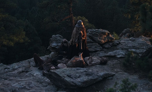 An armored figure stands over several people lying on a rocky ledge at dusk.