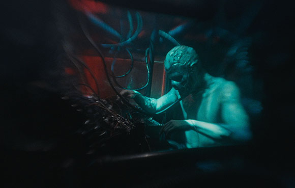 A pale, humanoid creature with textured skin reaches toward dark, tangled cables in a dimly lit setting.