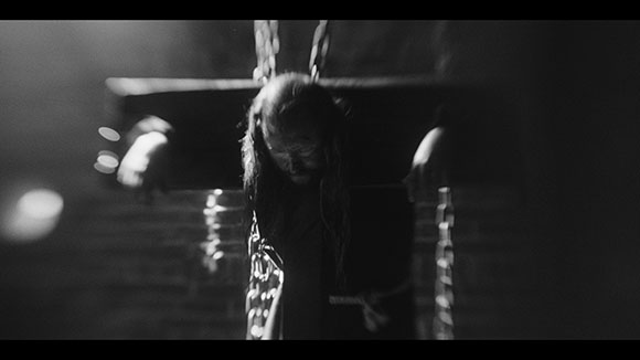Black and white shot of a person with long hair hanging from chains in a dark, dimly lit room.