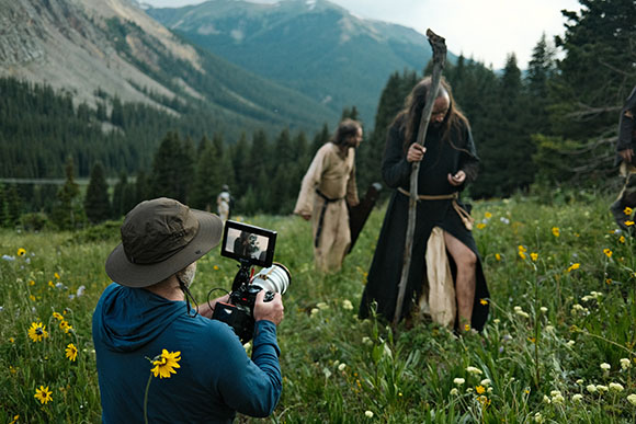 A photographer films two actors in long robes walking through a wildflower meadow in the mountains.