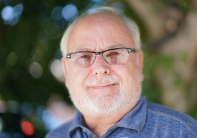A smiling older man with white hair, a white beard, and glasses wearing a blue shirt.