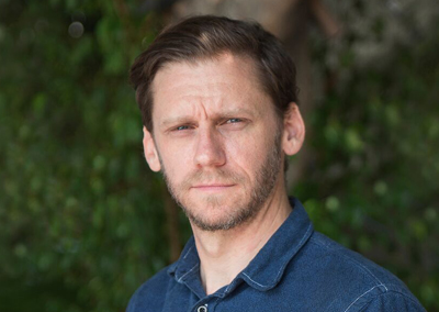 A man with short brown hair and a beard wearing a blue button-down shirt against a leafy green background.