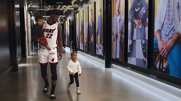 A man in a white Heat jersey carries a toddler while a young child walks next to him in a hallway with large portraits.