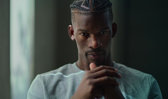 A man with short braided hair looks forward with his hands clasped near his chin.