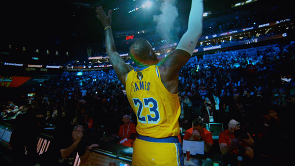 A basketball player in a yellow jersey with number 23 stands with arms raised facing a crowded arena.