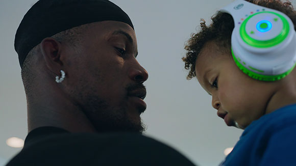A man in a black cap looks closely at a young child wearing white and green headphones.
