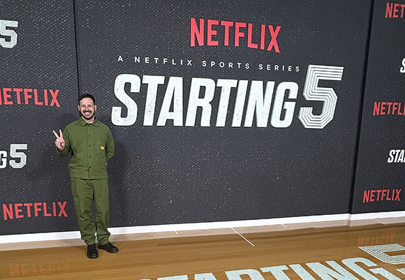 A man in an olive green jumpsuit makes a peace sign in front of a Netflix Starting 5 sports series backdrop.