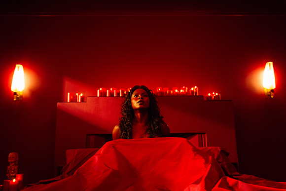 A woman with long curly hair sits in a bed under intense red lighting with candles in the background.