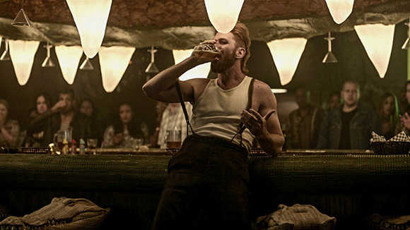 A bearded man in a white tank top and suspenders drinks from a glass at a dimly lit bar.