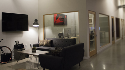 A lounge area with a dark sofa, coffee table, TV, and glass-walled offices in the background.