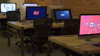 Multiple computer monitors on wooden desks with black office chairs in a dimly lit room.