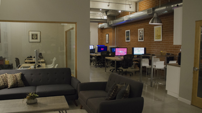 A lounge with grey sofas and a coffee table in the foreground, leading to a workspace with desks and computers.