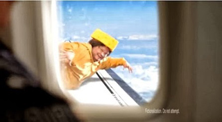 A person wearing a yellow suit and hat appears to be outside an airplane window against a cloudy sky.