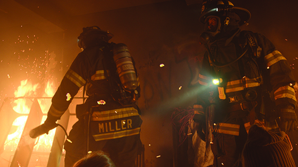 Two firefighters in full gear stand near a bright, intense fire with sparks flying in a dark environment.