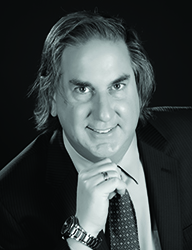 A man with medium-length hair wearing a dark suit and patterned tie, resting his chin on his hand.