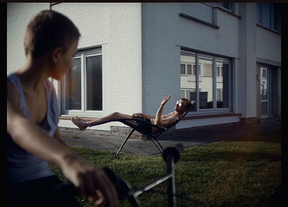 A person in a blue tank top looks toward someone reclining in a black lounge chair on a lawn.