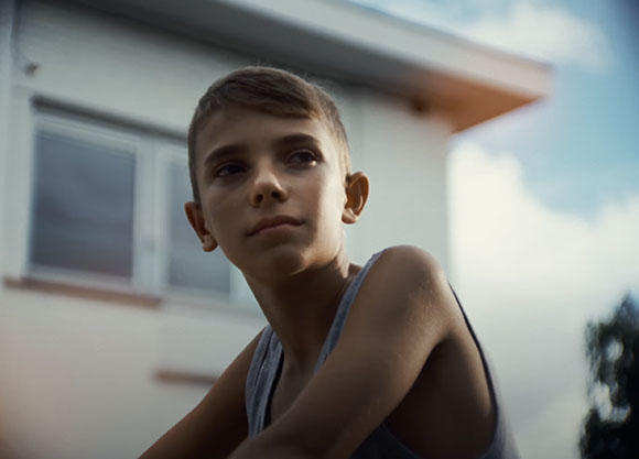 A young boy with short hair looking thoughtfully into the distance against a blurred house background.
