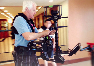 A man in a blue shirt uses a handheld camera stabilizer rig in a hallway.