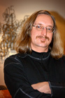 A man with long light brown hair, glasses, and a mustache poses with his arms crossed.