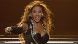 A woman with long wavy blonde hair smiling and wearing a black sequined outfit on stage.