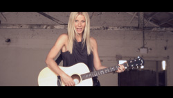 A blonde woman in a black sleeveless top playing an acoustic guitar in a rustic indoor setting.