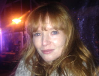 Close-up of a woman with long, wavy red hair and bangs, looking towards the viewer with a gentle smile. The background is blu