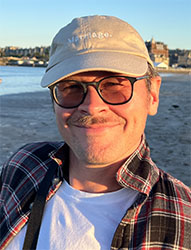 A man with a mustache, glasses, and a tan cap smiling outdoors near a beach.