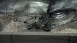 A massive, disc-shaped metallic object rests in a field of rubble and smoke under a cloudy sky.