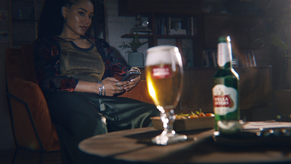 A woman sits in a chair using her phone near a glass of beer and a bottle on a table.