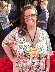 A smiling woman with glasses wearing a pink floral dress stands on a red carpet.