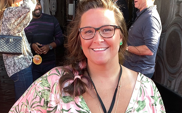 A smiling woman with glasses and a pink floral top stands in front of a crowd.
