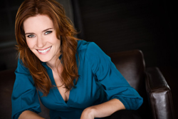 A woman with long, wavy red hair and fair skin smiles warmly. She is wearing a teal blouse and sitting in a dark leather chai