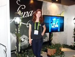 A woman with red hair stands in front of a display with a large screen showing a video and decorative plants.