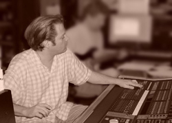 A man in a checkered shirt sits at a large audio mixing console in a studio.