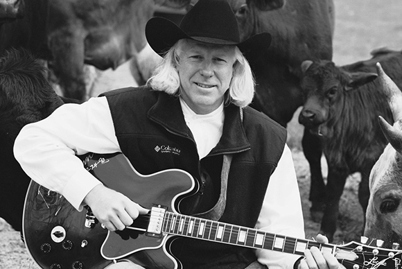 A man wearing a cowboy hat and vest holds a semi-hollow body electric guitar with cattle in the background.