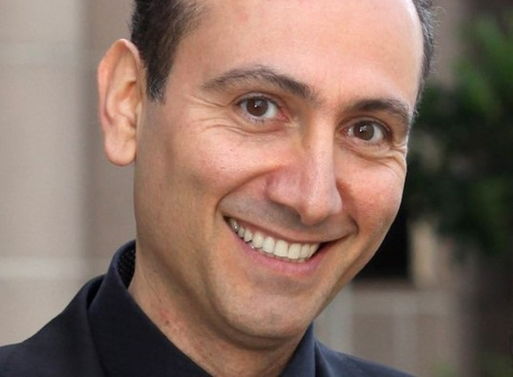A close-up portrait of a man with dark hair smiling at the camera.